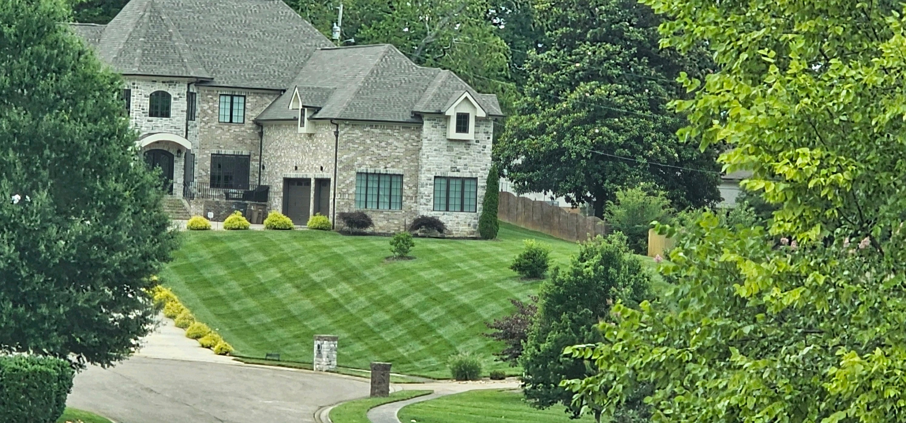 large gray brick house on a hill with a healthy green striped yard. lots of trees on the sides and background.