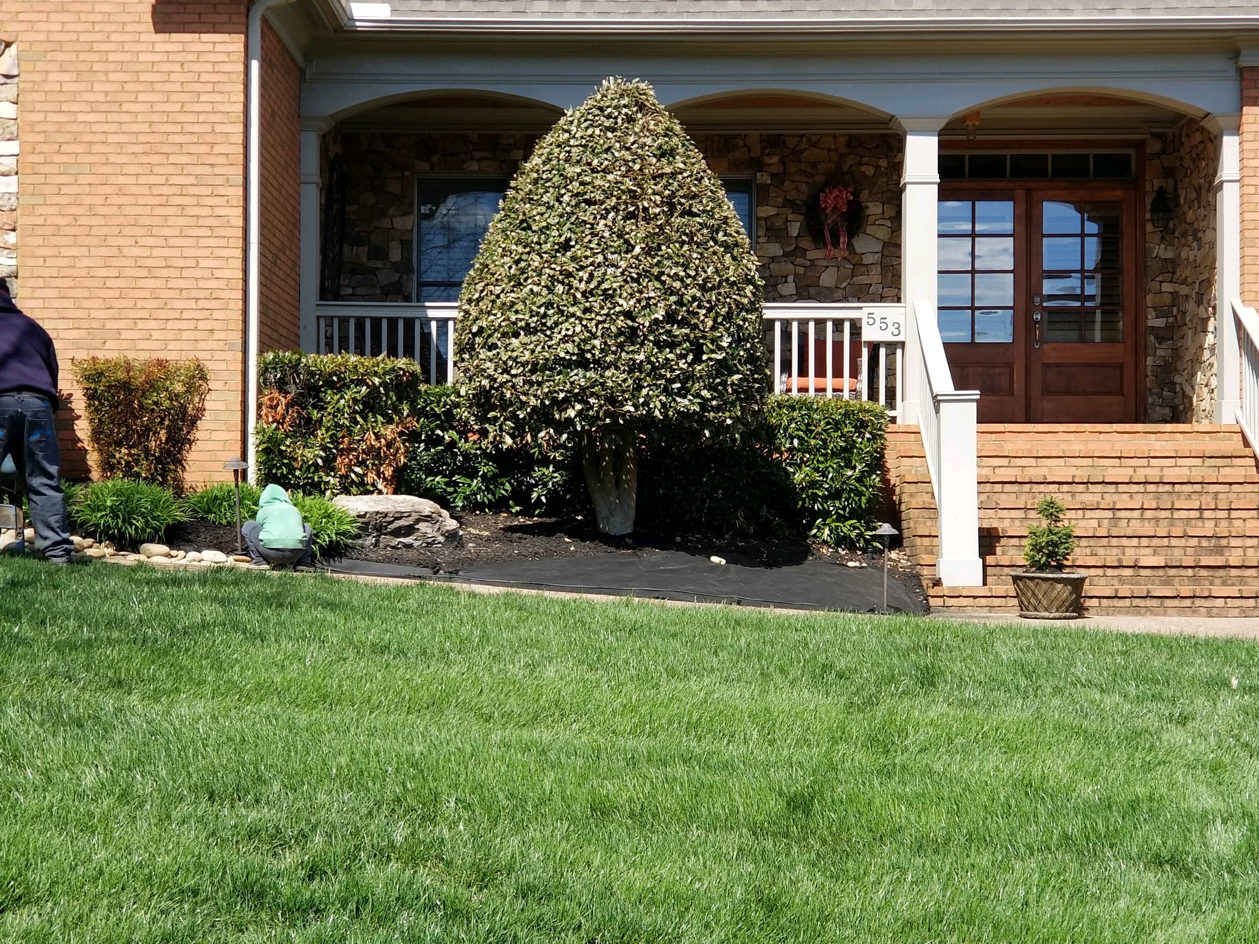 brick house with large porch. A perfectly trimmed bush in front of the house. green lawn in foreground.