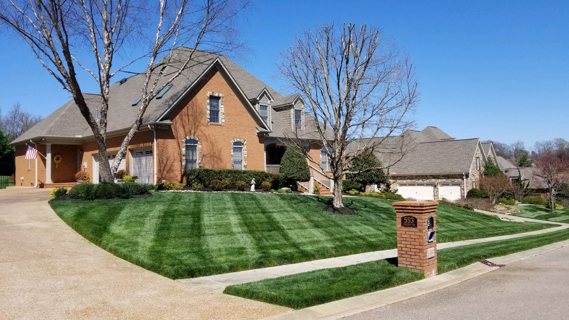 Brick home with healthy green striped yard. driveway on left and a few trees without leaves in the front yard.