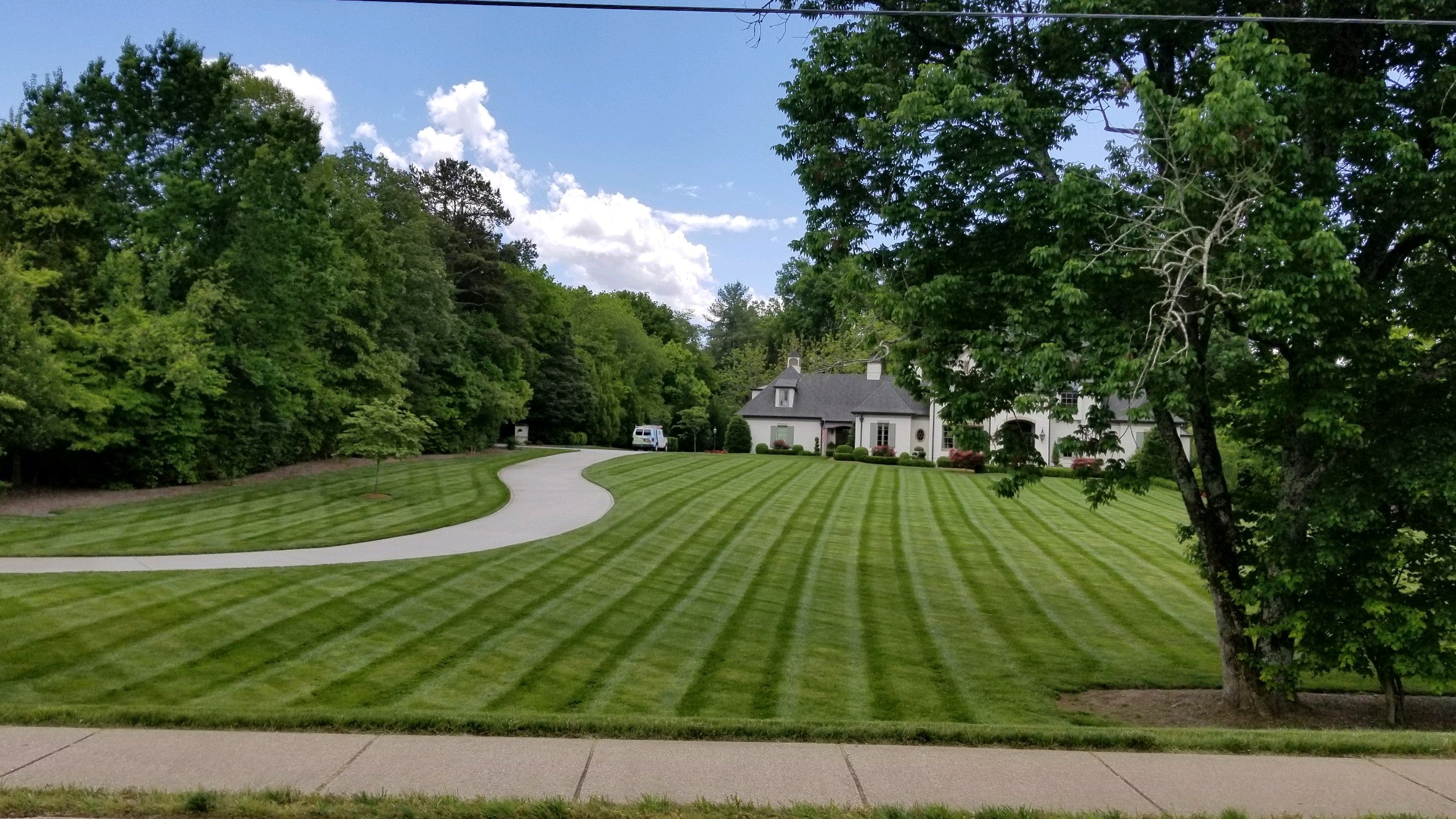 Large green striped lawn with driveway on left and large white brick home in back. Lots of trees in background behind teh house and on left side.