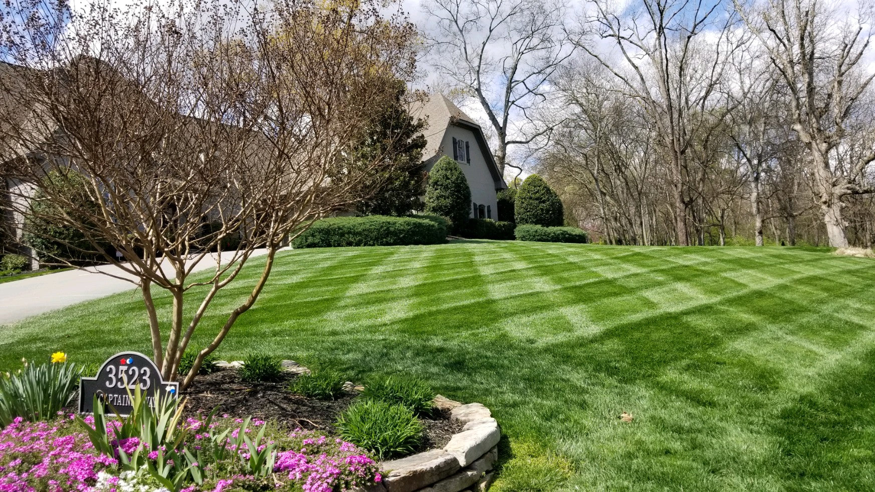 diagonal view of house in background with healthy green striped yard in foreground. small flowerbed on left front with tree and small plants. lots of trees behind house.