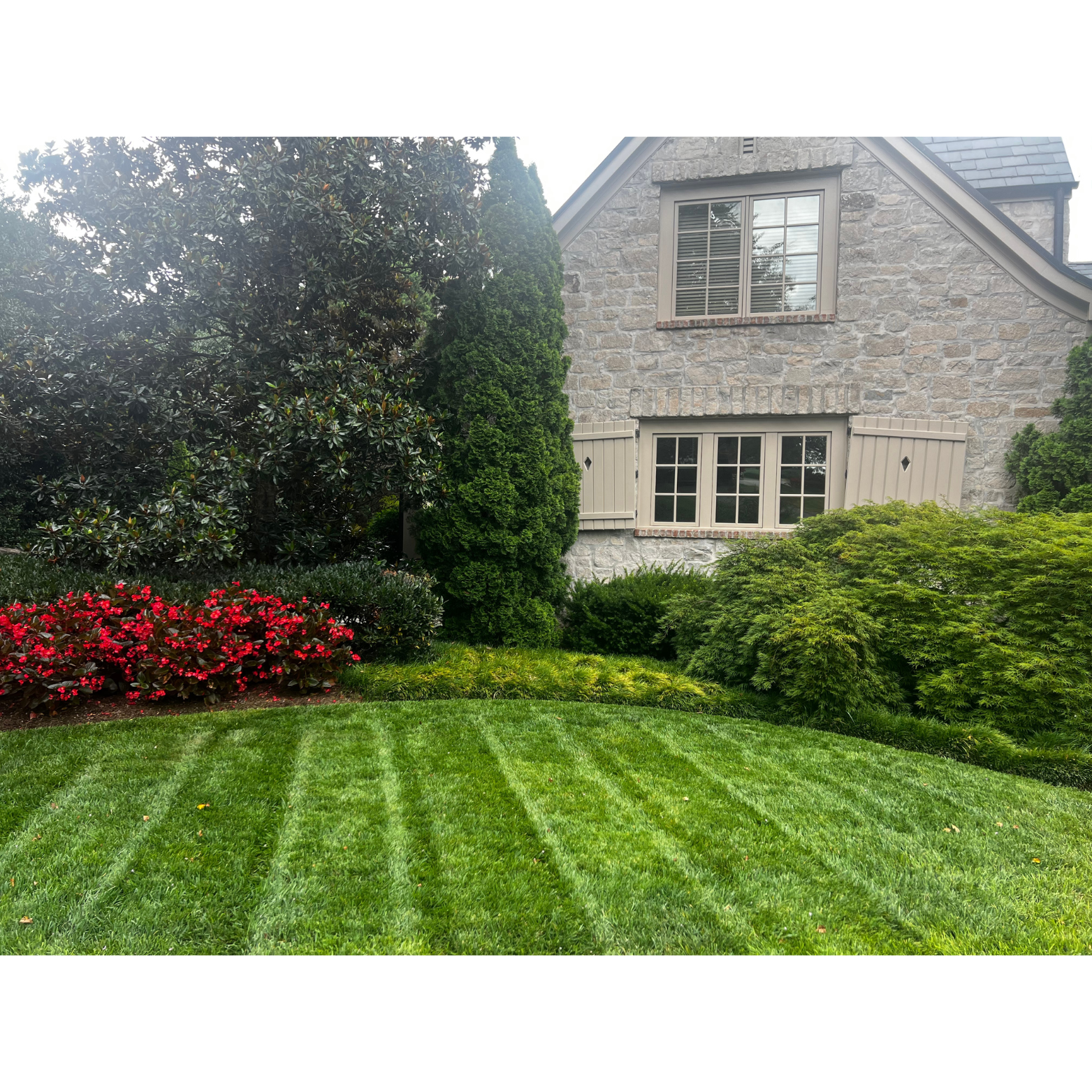 Gray Stone House with varius types of tree and plants with a pretty striped lawn.